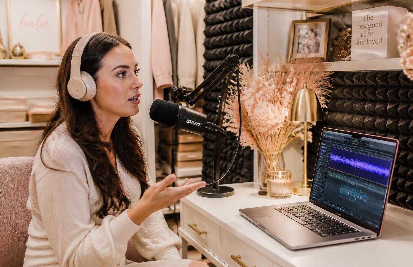 Woman recording a podcast episode in a cozy home closet studio with professional microphone and AI editing software on laptop